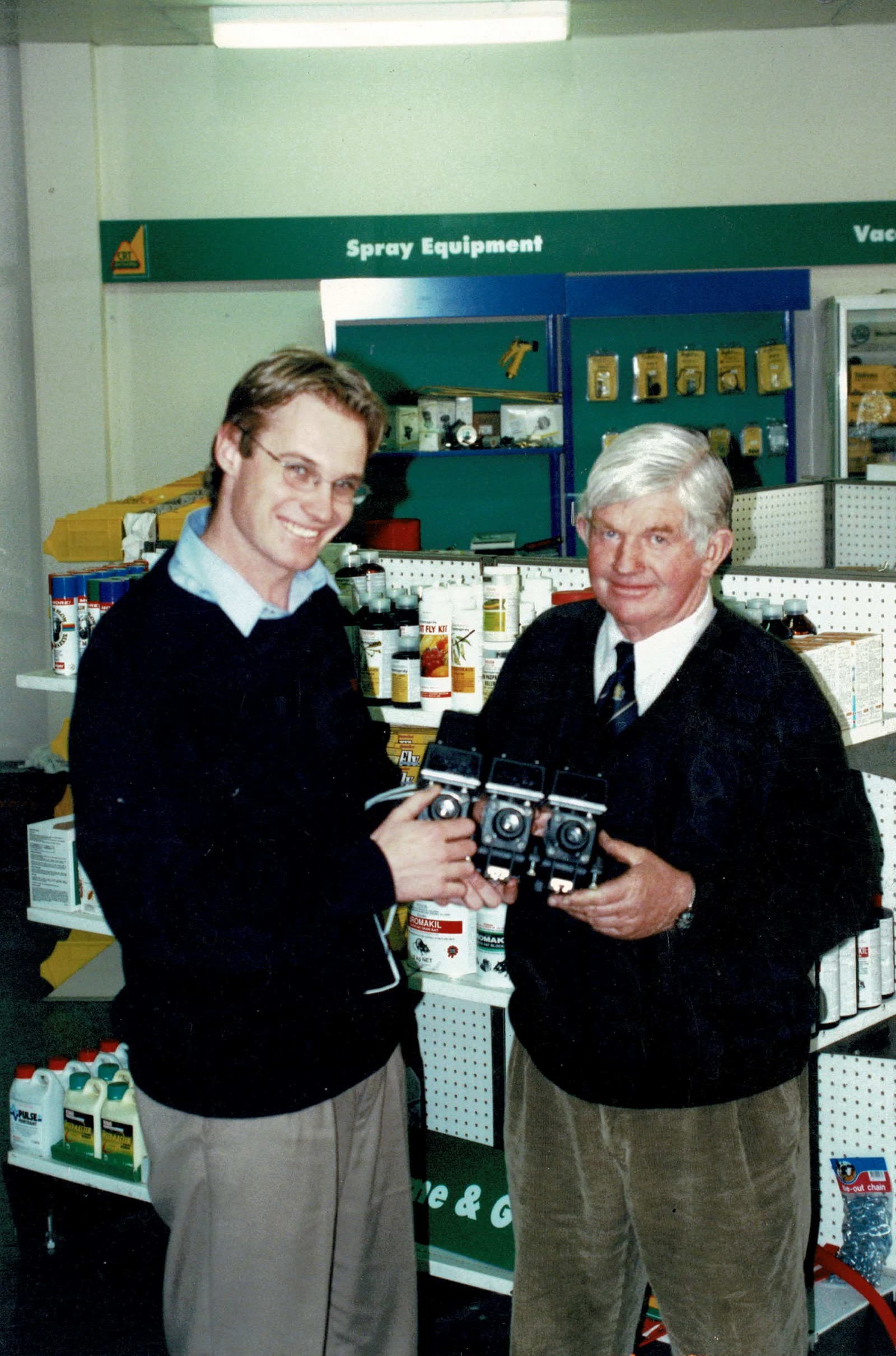 Two men in a rural supplies store holding a bank of three pumps under a spray equipment sign