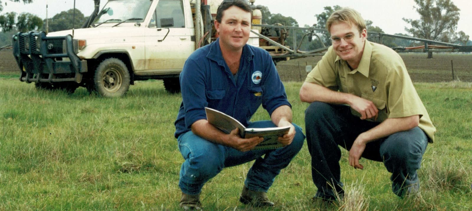 Two men crouched in a green paddock with a notebook, a ute and a spray unit behind them