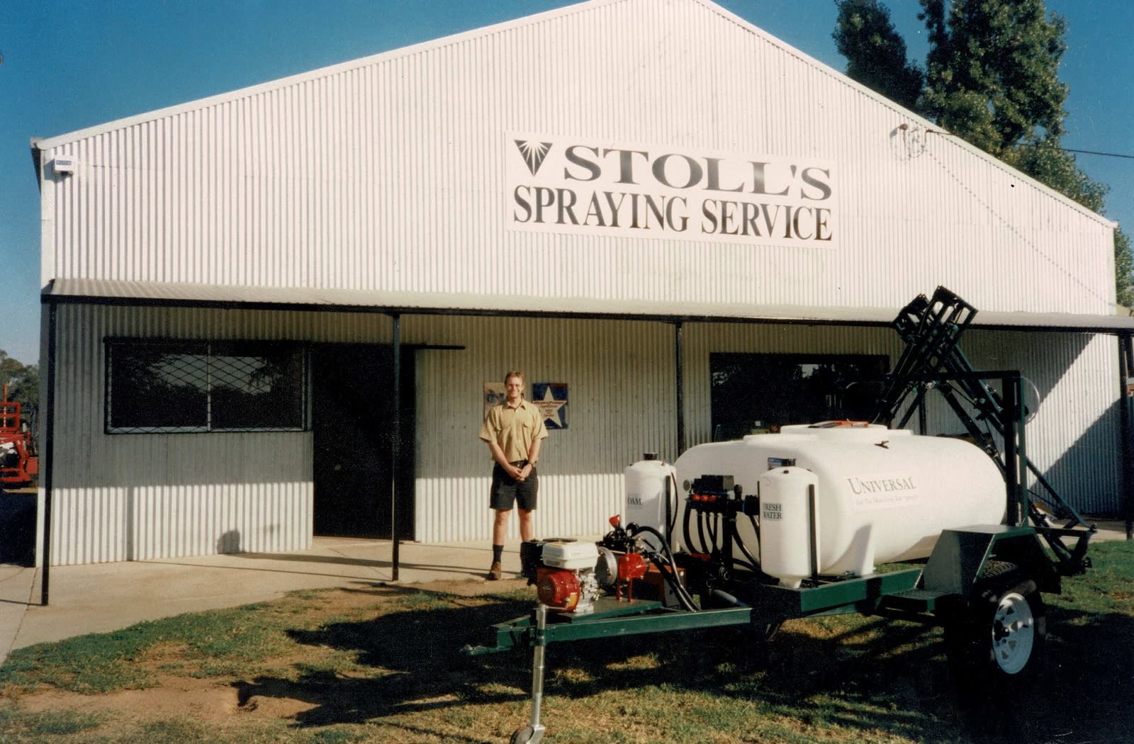 A man standing beside a small trailed spray unit outside a shed signed Stoll's Spraying Service